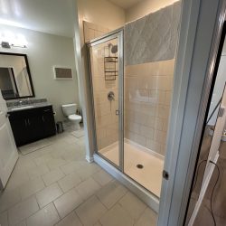 Modern bathroom with a glass shower enclosure, toilet, dark vanity, and tiled floor under bright overhead lighting.