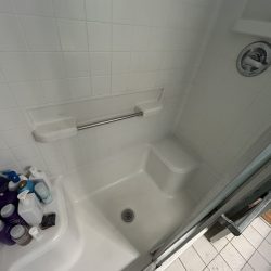 White tiled shower with a built-in seat, grab bar, assorted bottles, and a drain in the center of the floor.