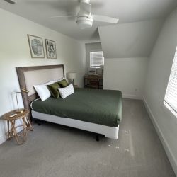 Minimalist bedroom with a green bedspread, two pillows, desk under window, ceiling fan, and neutral decor.