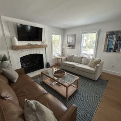 Living room with brown leather sofa, white loveseat, glass coffee table, fireplace, TV, and large windows.