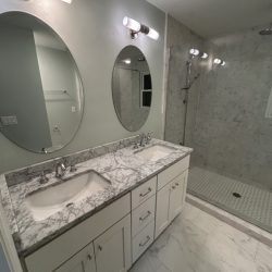 Modern bathroom with double sinks, marble countertop, oval mirrors, and a walk-in shower with glass door.