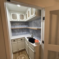 Small kitchenette with white cabinets, wood shelves, a sink, and a box on the counter; palm tree logo on the floor.