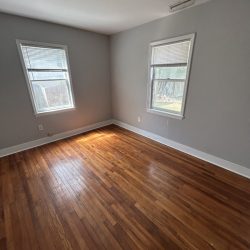 Empty room with two windows, light gray walls, wooden floor, and ceiling light fixture. Sunlight streams through one window.